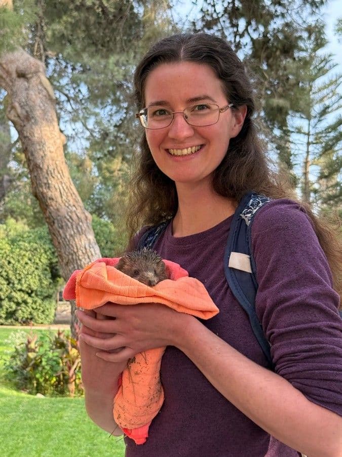 A smiling member of the JUC community holds a hedgehog wrapped in an orange towel outdoors.