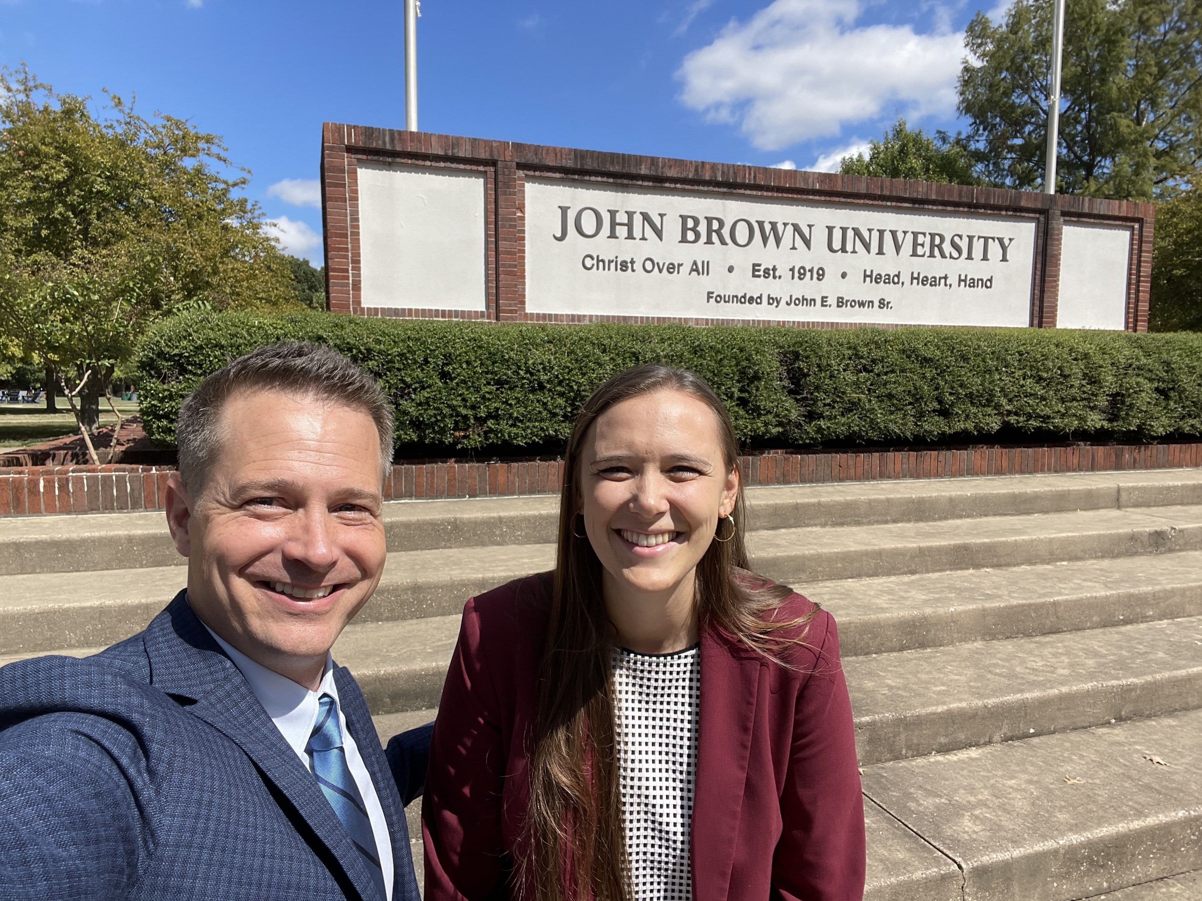 Dr. Oliver Hersey and Kaitlyn Hawn stand beside a John Brown University sign.
